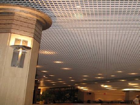 In a mall, the customers are walking and the lights shining on the steel grating ceiling.