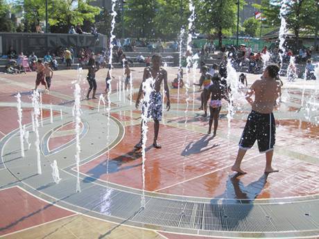 Sur un carré, la fontaine pulvérise de l'eau sur un râlage en acier façonné et les enfants jouent.